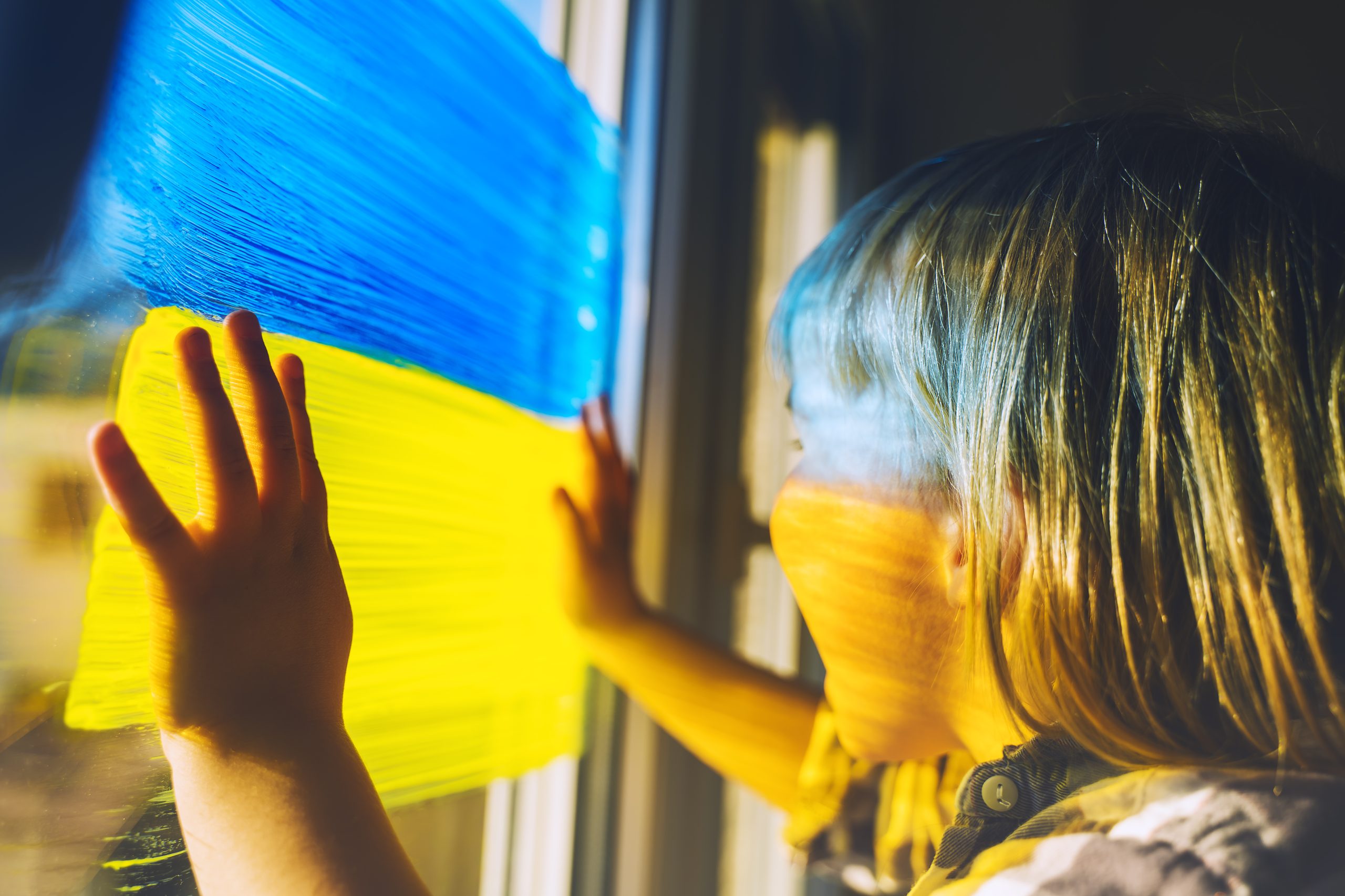 Little child with hands on yellow-blue flag image of Ukraine on window. Support Ukrainians. Concept, emotions. Color symbol image of flag of Ukraine on glass with paints.